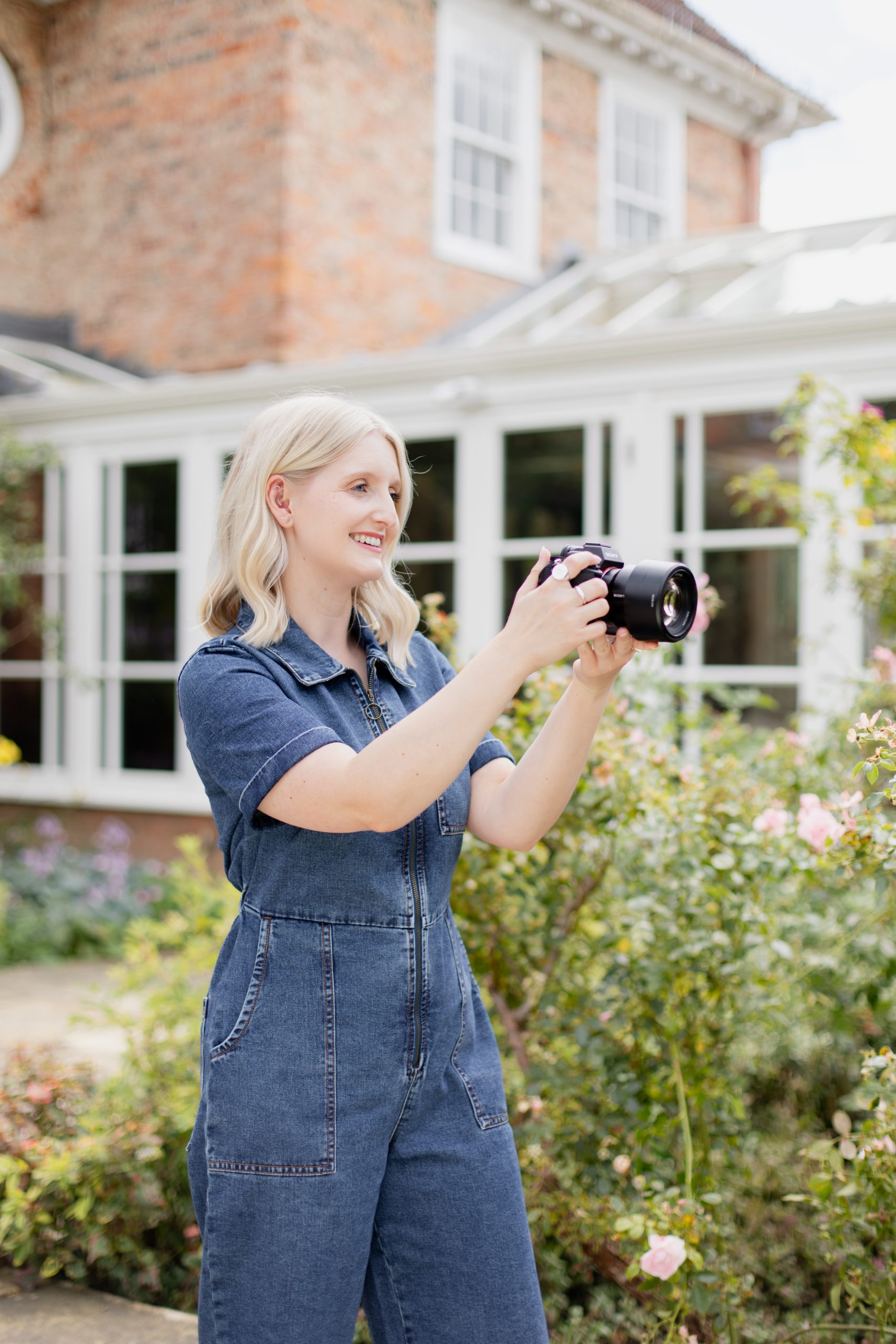 candid wedding photographer Beth Lavin holding camera outside wearing a blue denim jumpsuit